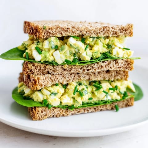 Creamy Avocado Egg Salad Sandwich served on whole grain bread, garnished with chives and a lemon wedge for brightness.