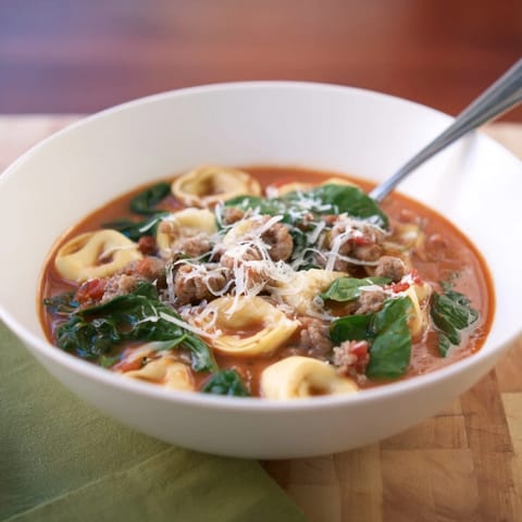 Creamy Tuscan Tortellini Soup ladled into a white bowl, topped with grated Parmesan and fresh basil on a cozy table. 